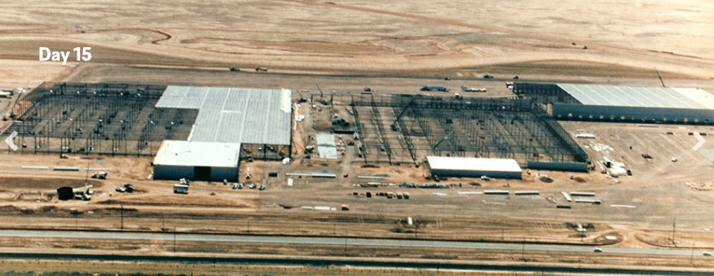 Aerial view labeled “Day 15” showing rapid construction progress, with large steel framing erected, partial roof panels installed, and multiple building sections taking shape across a desert job site.